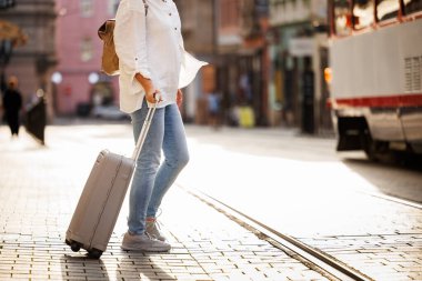 Kadın turist bavulunu çekiyor ve şehirde tramvay raylarıyla caddede yürüyor. Seyahat ve tatil yaşam tarzı