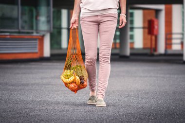 Tekrar kullanılabilir ağ çantalı bir kadın süpermarketin yanındaki otoparkta yürüyor. Plastik ücretsiz alışveriş ve sıfır atık konsepti. Meyve ve sebze alışverişi.