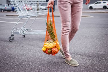 Tekrar kullanılabilir ağ çantalı bir kadın süpermarketin yanındaki otoparkta alışveriş arabasından yürüyor. Plastiksiz ve sıfır atık konsepti. Market alışverişi