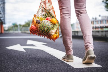 Sıfır atık ile sağlıklı ve sürdürülebilir yaşam tarzı için doğru yönü seçin. Elinde meyve dolu çanta olan bir kadın süpermarketten yolda yürüyor.