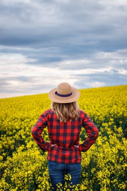 Gururlu çiftçi gün batımında çiçek açan yağ tohumu tarlasına bakıyor. Hasır şapkalı ve ekose gömlekli kadın kolza tohumu tarlasında duruyor.