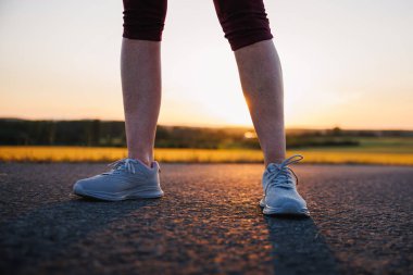 Gün batımında koşu ayakkabılı bir kadın yolda duruyor. Açık havada spor eğitimi.