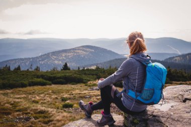 Dağ yürüyüşü sırasında rahatlama. Sırt çantalı bir kadın kayanın üzerinde oturuyor ve manzaranın tadını çıkarıyor. Dağ zirvesinde turist yürüyüşü ve tırmanışı