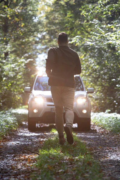 man running to car standing at the forest trail with headlights turned on