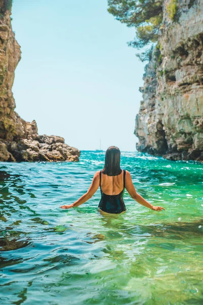 身穿黑色泳衣的年轻成年女子走在岩石悬崖之间的海面上夏季海滩度假 — 图库照片