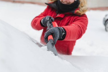 Kış mevsimi kar fırtınasından sonra kürk kapüşonlu bir adam araba temizliyor.