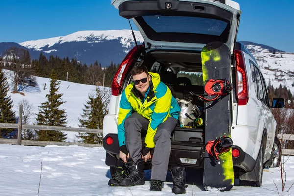 man sitting in car trunk changing for snowboard. mountains on background