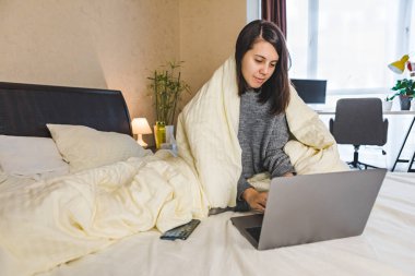 Yatakta çalışan bir kadın, battaniye ile kaplı bir dizüstü bilgisayarla çalışıyor.