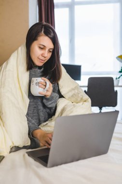 Yatakta dizüstü bilgisayarda çalışan ve battaniyeli kahve içen bir kadın.