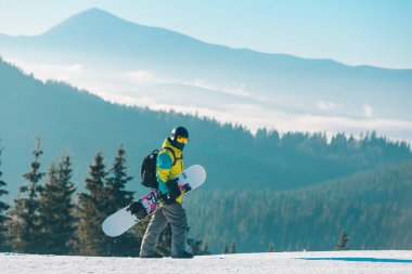 Arka planda snowboard dağlarıyla tepenin yanından geçen bir adam.