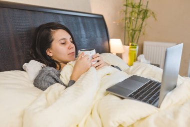 Kadın yatakta kahve içiyor ve dizüstü bilgisayarla çalışıyor.