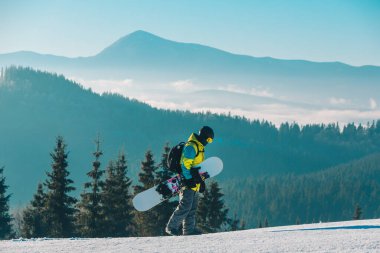 Arka planda snowboard dağlarıyla tepenin yanından geçen bir adam.