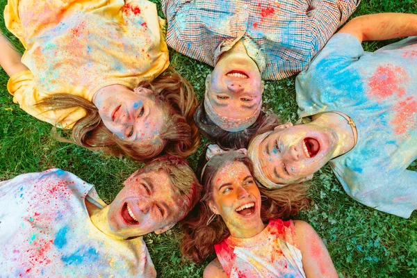 Amigos felizes que colocam no chão grama verde sujo em tintas holi — Fotografia de Stock