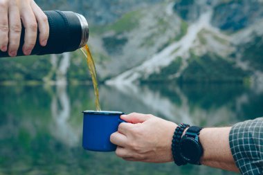 Termostan fincana çay doldurmak konsept yürüyüş gölü ve arka planda dağlar ısınma içkisi