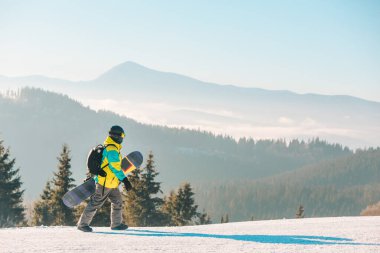 Arka planda snowboard dağlarıyla tepenin yanından geçen bir adam.