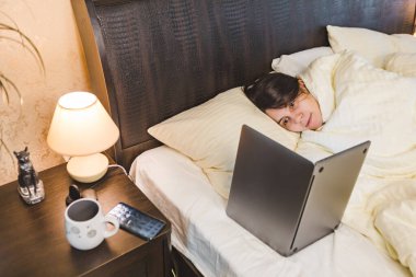 Kadın battaniyeyle kaplı bir şekilde yatağa uzanmış dizüstü bilgisayarın fotokopisini çekiyor.