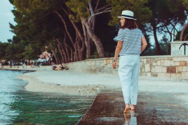 Beyaz elbiseli ve fötr şapkalı bir kadın yaz mevsiminde deniz kıyısında yürüyor.