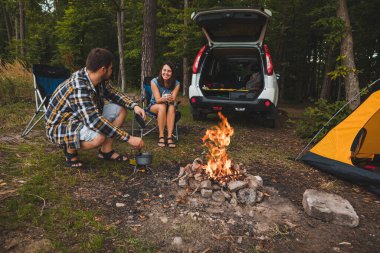 Kamp ateşinin yanında oturan çift. Adam ateşte pişiyor. Yürüyüş konsepti. araba yolculuğu