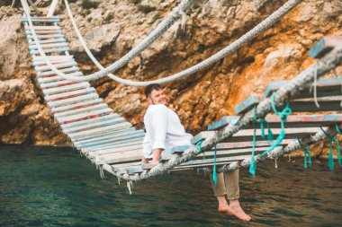 Asma köprüde oturan adam deniz manzarası ve doğal sakinliğin tadını çıkarıyor.