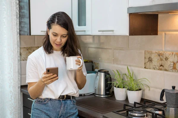 Woman drinking morning coffee tea at the kitchen surfing internet on the phone — Stock Photo, Image