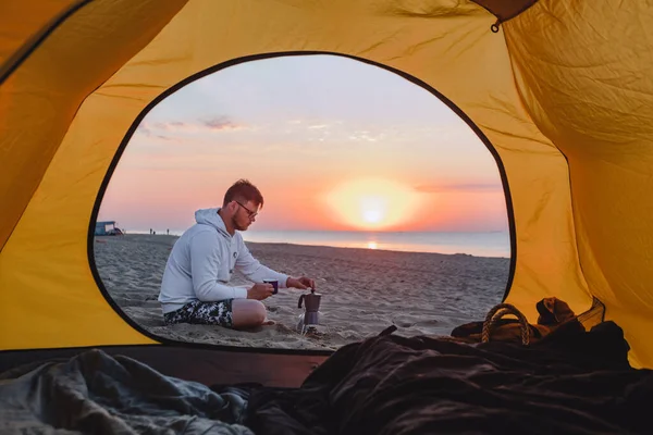 男が海の砂浜でコーヒーを作ってる 夏休みキャンプ テントを通して見る — ストック写真