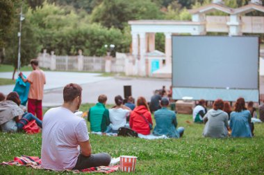 Şehir parkındaki açık hava sinemasında battaniyenin üzerinde oturan adam büyük ekranın önünde abur cubur yiyor.
