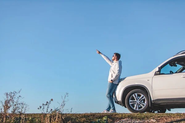 Mladý dospělý muž stojící poblíž auta v slunečních brýlích modrá obloha na pozadí — Stock fotografie