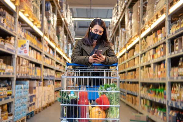 Kobieta Sprawdzanie Telefonu Podczas Robienia Zakupów Spożywczych Masce Medycznej Przestrzeń — Zdjęcie stockowe
