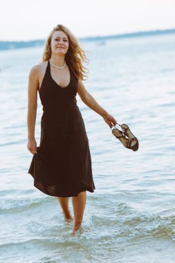 Siyah açık renk elbiseli bir kadın ılık yaz akşamında deniz kenarında yalınayak yürüyor.