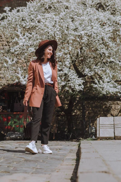 Улыбающаяся Красивая Женщина Куртке Шляпе Прогуливаясь Весенней Улице Белого Цветущего — стоковое фото