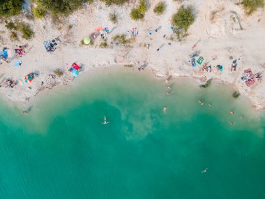 Mavi gök mavisi suda yüzen, kumlu sahilde güneşlenen insanların havadan görüntüsü.