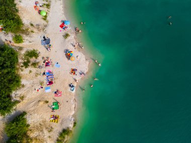 Mavi gök mavisi suda yüzen, kumlu sahilde güneşlenen insanların havadan görüntüsü.