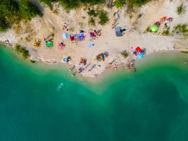 Mavi gök mavisi suda yüzen, kumlu sahilde güneşlenen insanların havadan görüntüsü.