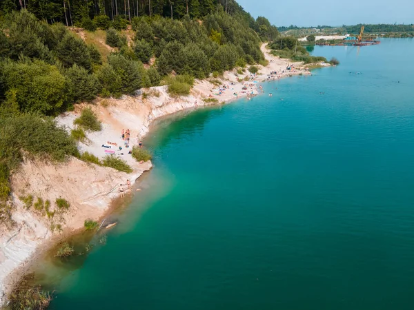 Mavi gök mavisi suda yüzen, kumlu sahilde güneşlenen insanların havadan görüntüsü.