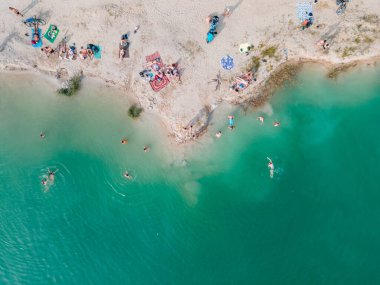 Mavi gök mavisi suda yüzen, kumlu sahilde güneşlenen insanların havadan görüntüsü.