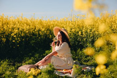 Beyaz elbiseli çekici bir kadın battaniyeye oturmuş şarap içiyor.