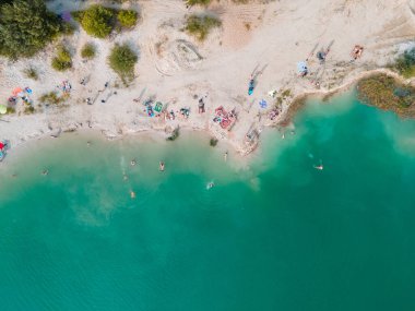 Mavi gök mavisi suda yüzen, kumlu sahilde güneşlenen insanların havadan görüntüsü.