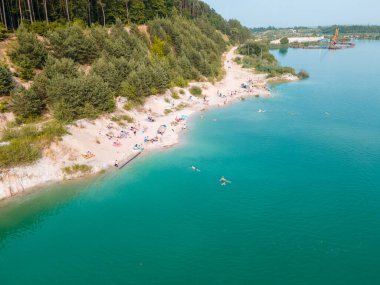 Mavi gök mavisi suda yüzen, kumlu sahilde güneşlenen insanların havadan görüntüsü.