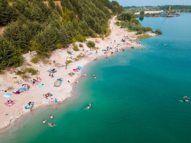 Mavi gök mavisi suda yüzen, kumlu sahilde güneşlenen insanların havadan görüntüsü.