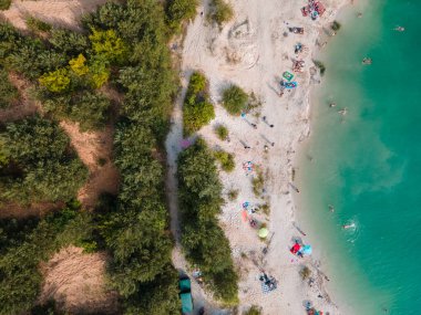 Mavi gök mavisi suda yüzen, kumlu sahilde güneşlenen insanların havadan görüntüsü.