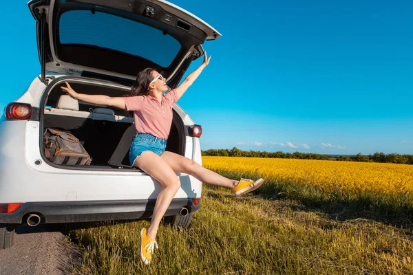 Lächelnde hübsche Frau sitzt im Kofferraum eines Geländewagens bei Sonnenuntergang und blickt auf ein Feld — Stockfoto