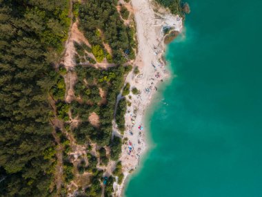 Mavi gök mavisi suda yüzen, kumlu sahilde güneşlenen insanların havadan görüntüsü.