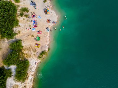 Mavi gök mavisi suda yüzen, kumlu sahilde güneşlenen insanların havadan görüntüsü.