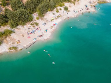 Mavi gök mavisi suda yüzen, kumlu sahilde güneşlenen insanların havadan görüntüsü.