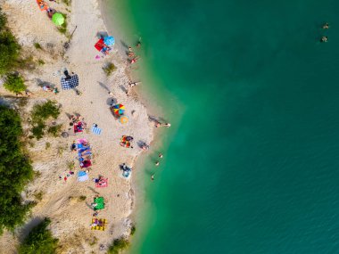 Mavi gök mavisi suda yüzen, kumlu sahilde güneşlenen insanların havadan görüntüsü.