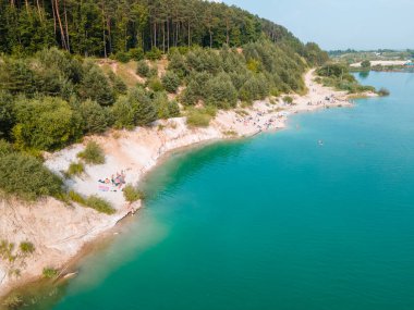 Mavi gök mavisi suda yüzen, kumlu sahilde güneşlenen insanların havadan görüntüsü.
