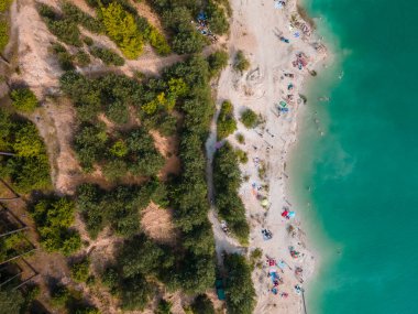 Mavi gök mavisi suda yüzen, kumlu sahilde güneşlenen insanların havadan görüntüsü.