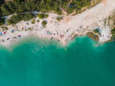 Mavi gök mavisi suda yüzen, kumlu sahilde güneşlenen insanların havadan görüntüsü.
