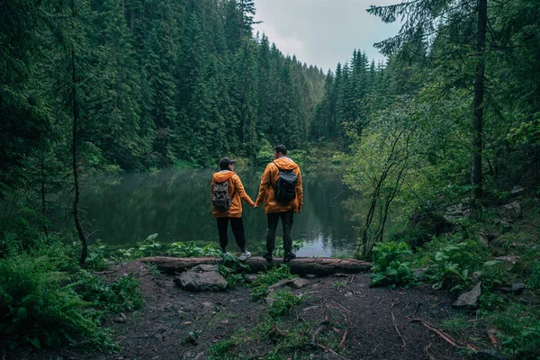 Casal caminhantes em capa de chuva amarela olhando para lago de montanha — Fotografia de Stock
