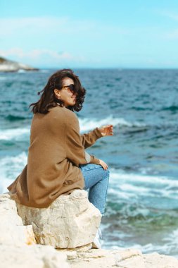 uçurumun kenarında oturup deniz manzarasının keyfini çıkaran kadın. rüzgarlı hava. güneşli bir gün. yaz tatili. kopyalama alanı
